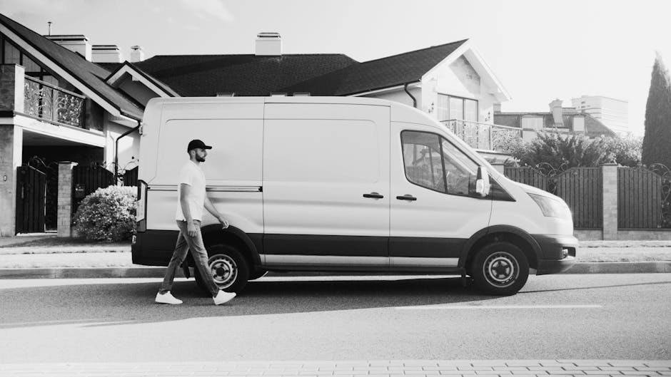 A man wearing a cap, t-shirt, and jeans is walking alongside a white panel van parked on the street outside a residential property. The van is positioned parallel to the pavement, with its side door closed, and the vehicle appears to be used for home relocation or furniture transport. The background features a modern house with a tiled roof, a balcony, and a garden area enclosed by a fence. The environment is well-lit, indicating daytime, and the scene captures the loading process as part of a house removals service, with the van ready for the next stage of packing or transporting furniture and boxes, supported by the professional removal company, Man with Van Colliers Wood.