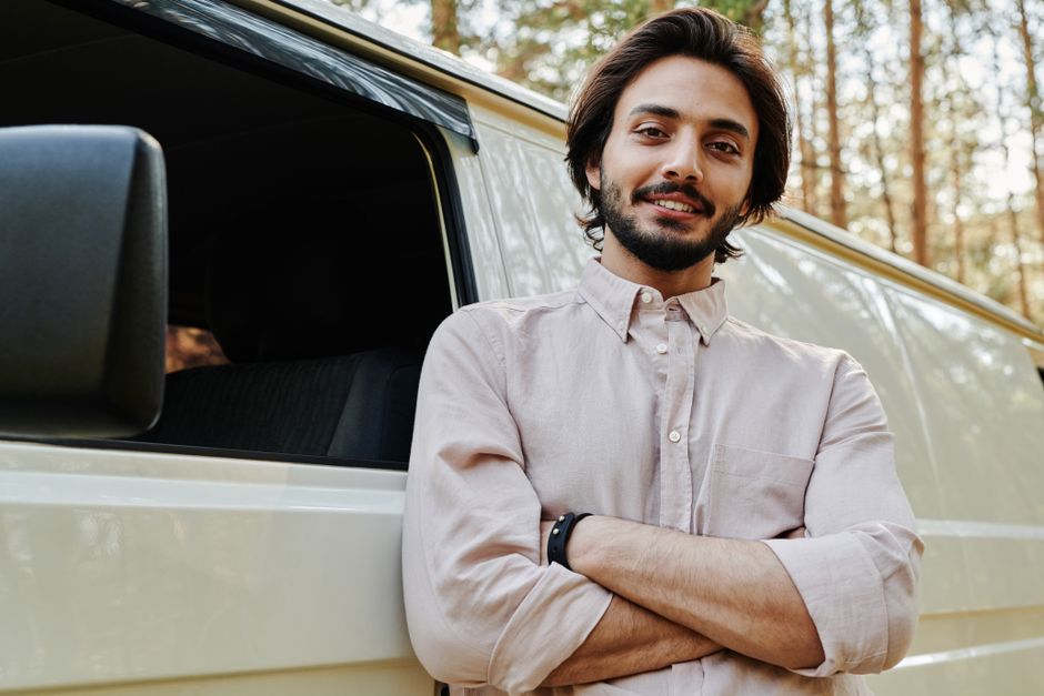 A young man with dark, shoulder-length hair and a beard, standing outdoors beside a white moving van with a black side mirror, slightly smiling with arms crossed, wearing a light beige button-up shirt and a black wristwatch. The background features a wooded area with trees and sunlight filtering through the leaves, indicating daylight. The vehicle is positioned on a paved surface, and the scene suggests preparation or completion of a home relocation or furniture transport process, with the van likely used for packing and moving household items as part of professional removals services offered by Man with Van Colliers Wood.