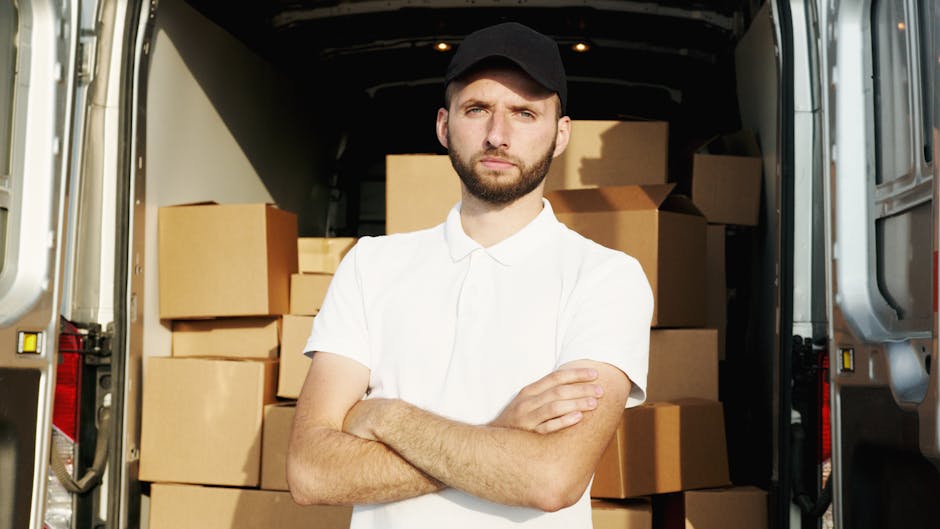 A young man with a beard, wearing a black cap and a white polo shirt, stands inside the open rear of a moving van filled with stacked cardboard boxes of various sizes, some wrapped in plastic or fabric for protection. The van is parked outdoors on a pavement near a doorway, with natural daylight illuminating the scene. The man has his arms crossed and a serious expression, indicating involvement in a home relocation or furniture transport process. The boxes are arranged for efficient loading or unloading, and the interior of the van appears prepared for moving services offered by Man with Van Colliers Wood, as shown on the page about furniture removals in Colliers Wood.