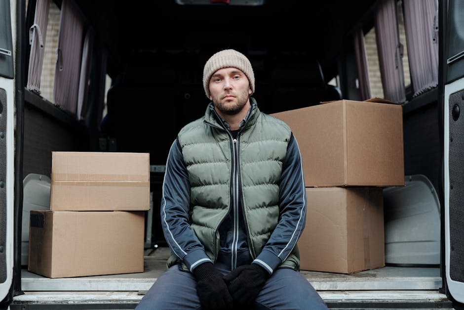 A man wearing a beige knit beanie, green puffer vest, and navy blue tracksuit top is sitting on the edge of an open van's loading area, surrounded by several cardboard moving boxes of varying sizes. The boxes are sealed with packing tape, with some stacked on top of each other and others placed directly on the van floor. The van's interior is dark, with the rear doors open to reveal a driveway or street outside, indicating the loading process during a house relocation. The man appears to be taking a break or waiting during the packing and moving operation managed by Man with Van Colliers Wood, whose company name is subtly referenced. The scene captures a moment of furniture transport and packing logistics, with the focus on the boxes and the loading area, illustrating the process of home relocation involving careful handling and transportation of personal belongings.