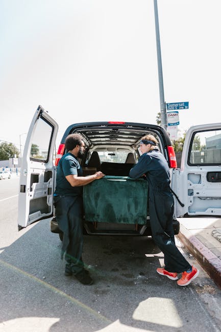 Two professional movers from Man with Van Colliers Wood are loading a large, heavy object covered with a green moving blanket into the open rear of a black van on a city street. The van is parked on the side of the road, with its rear doors wide open, revealing the interior space used for packing and transporting furniture and bulky items. The movers are wearing dark uniforms, with one in a short-sleeved shirt and the other in a long-sleeved jacket, both focused on carefully lifting the item to ensure safe handling during the home relocation process. In the background, a lamppost with a blue street sign indicating Wandle Park is visible, along with clear skies and some distant buildings, suggesting a typical urban environment for furniture transport and packing during a house move. The scene captures the loading process essential for professional removals services by [COMPANY_NAME], highlighting the equipment such as moving blankets used for protection and the logistical steps involved in house removals and furniture transport.
