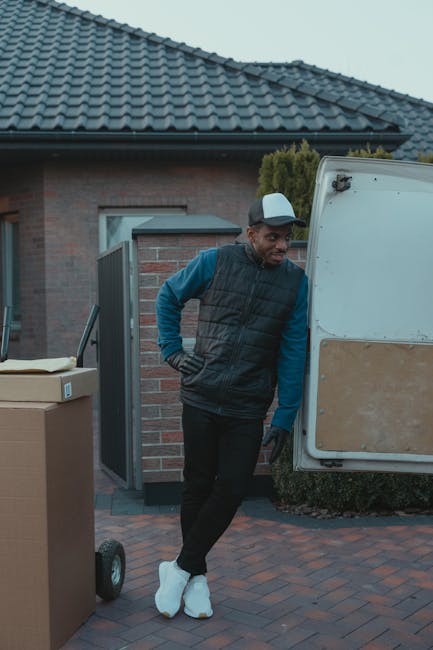 A man wearing a beige knit beanie, green puffer vest, and navy blue tracksuit top is sitting on the edge of an open van's loading area, surrounded by several cardboard moving boxes of varying sizes. The boxes are sealed with packing tape, with some stacked on top of each other and others placed directly on the van floor. The van's interior is dark, with the rear doors open to reveal a driveway or street outside, indicating the loading process during a house relocation. The man appears to be taking a break or waiting during the packing and moving operation managed by Man with Van Colliers Wood, whose company name is subtly referenced. The scene captures a moment of furniture transport and packing logistics, with the focus on the boxes and the loading area, illustrating the process of home relocation involving careful handling and transportation of personal belongings.